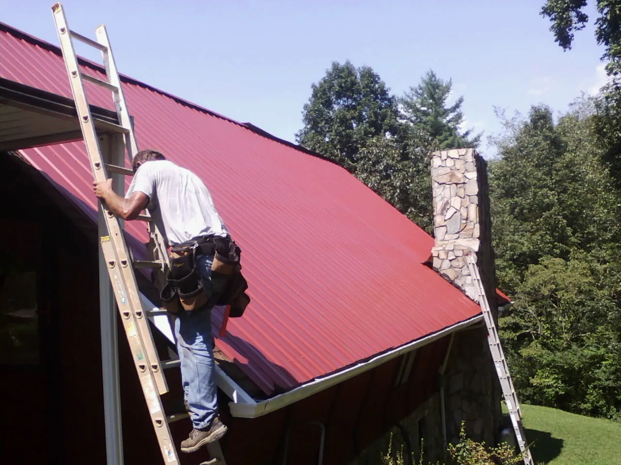 Roofer on a ladder beside a red metal roof and stone chimney — Soffit & Fascia Repair by Roofing Brothers in Letha